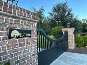 Elegant black gate with a brick pillar marked 'BENTWOOD', surrounded by greenery and a clear sky.